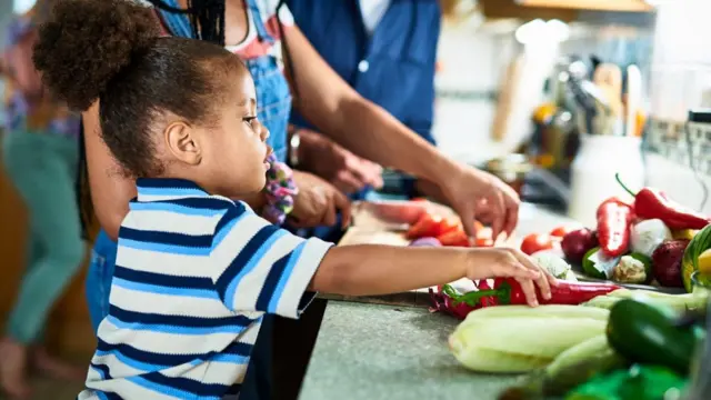 Un niño pequeño con el cabello recogido toca un pimiento rojo sobre el mesón de una cocina, mientras una mujer corta verduras a su lado.