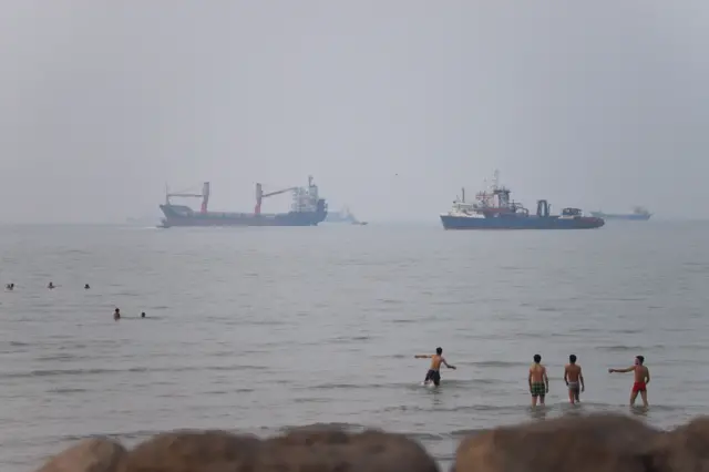 Buques de carga y petroleros permanecen en primer plano frente a la costa, mientras que personas nadan y se refrescan en las aguas costeras en un día brumoso.
