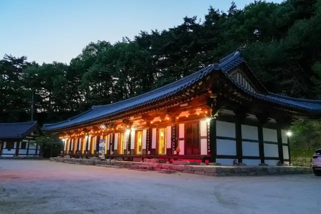 Templo tradicional coreano iluminado con luces cálidas al atardecer, situado en un tranquilo patio al borde de una ladera boscosa.