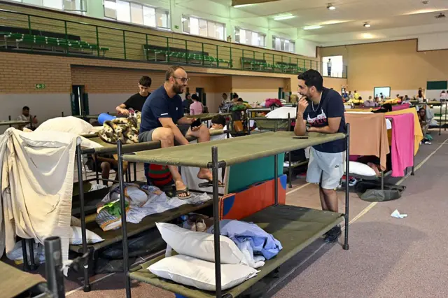Literas de campaña hechas con tubos y lona de color militar se alienean en un polideportivo. La gente ha puesto en ellas sus almohadas, sábanas o ha colgado sus toallas. En una de ellas, sentado en la parte de arriba, hay un hombre con pantalón corto y camiseta de manga corta que conversa con otro, que está apoyado en otra de las literas y también viste pantalón corto y camiseta de manga corta. Al fondo se ve más gente. 