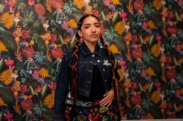 Renata Flores vestida con una chaqueta de jeans, un fajín negro lleno de bordados de colores, dos trenzas en el pelo, largas, hasta la cintura, adornadas con lanas de colores. El fondo de la pared es una papel pintado llenos de motivos florales.