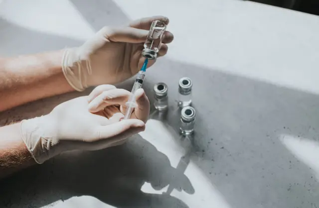 Una mano con guantes de látex sostiene un vial de líquido transparente y una jeringa