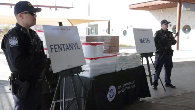 Una mesa repleta de paquetes de polvo blanco y dos letreros que dicen fentanilo y metanfetamina. Dos hombres uniformados con gorras de béisbol montan guardia. 