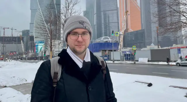 En primer plano, una persona con gorro gris, gafas, abrigo negro y bufanda blanca lleva una mochila gris con las correas visibles.