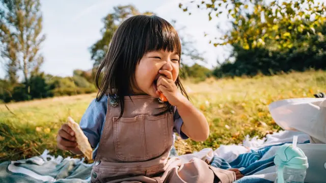 Una niña pequeña sentada al aire libre come una zanahoria mientras sostiene un trozo de pan en la otra mano.
