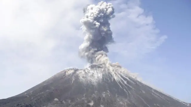 Fotografía de un volcán activo que emite una columna de ceniza y vapor desde su cima sobre un paisaje costero.