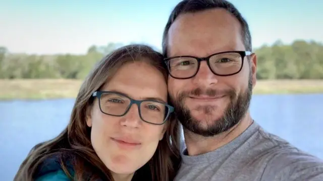 Katie y Alan Donegan sonríen a la cámara mientras ambos llevan gafas, tomándose una selfi frente a un lago y árboles bajo un cielo azul.