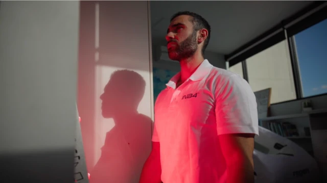 Lucas está de pie en el interior de una casa, con una luz roja proyectada sobre su cuerpo. Tiene el pelo corto y oscuro, y barba oscura. Viste una camiseta blanca de cuello alto.