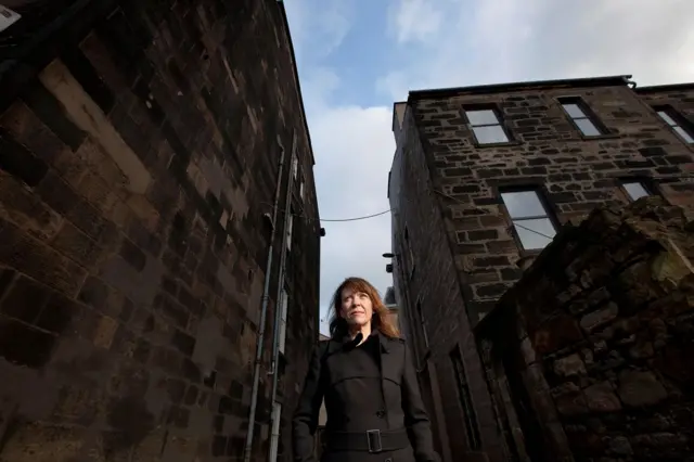 Karyn McCluskey, una mujer pelirroja con una gabardina negra, vista en contrapicado en medio de dos casas en Glasgow.
