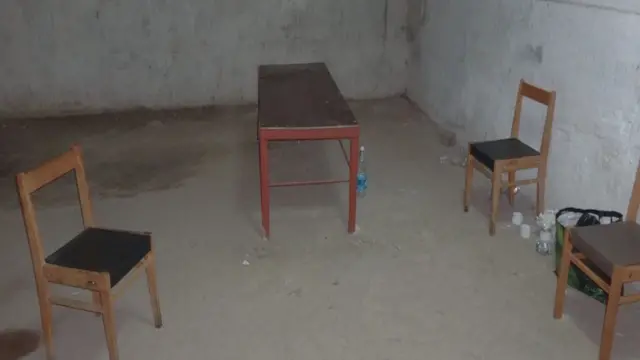 Una habitación desnuda, con paredes y suelo de hormigón. En ella hay tres sillas de madera y una pequeña mesa de madera, con unas cuantas botellas de agua de plástico en el suelo.