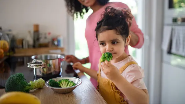 Una niña pequeña está de pie junto al mesón de una cocina mientras huele un trozo de brócoli.