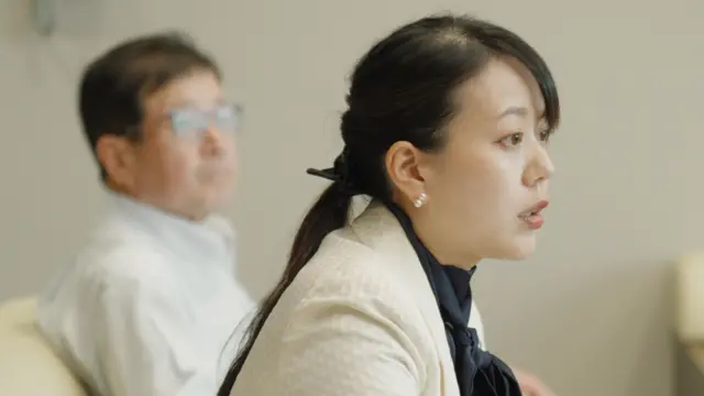 Shoko Kawata, una alcaldesa vestida con una chaqueta blanca y una blusa oscura, está sentada en un sillón. Detrás de ella se ve a su adjunto, que lleva una camisa de color claro.
