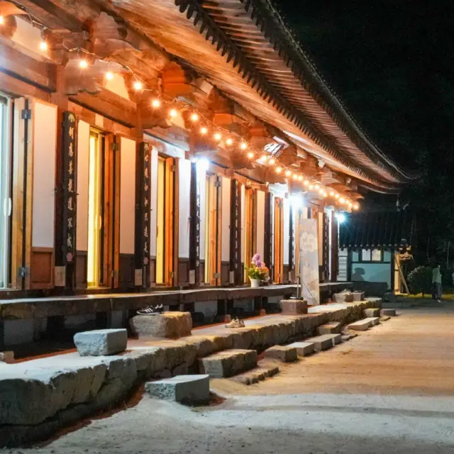 Un templo coreano tradicional iluminado por cálidas luces por la noche, con un tejado curvo de tejas, columnas de madera y escalones de piedra que bordean un amplio patio. Varios visitantes caminan por el sendero que discurre junto al templo.