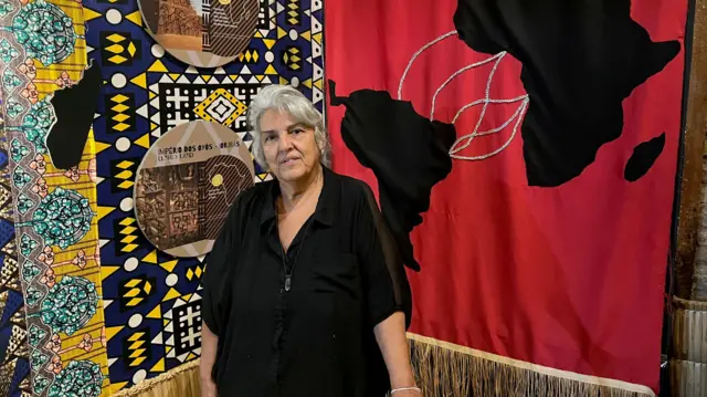 Una mujer de cabello gris, de pie. Detrás de ella hay un mural que muestra un dibujo del mapa de África conectado con América del Sur.