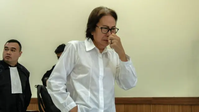 Una mujer con una camisa blanca y gafas de montura negra parece emocionada y se lleva un pañuelo a la nariz.