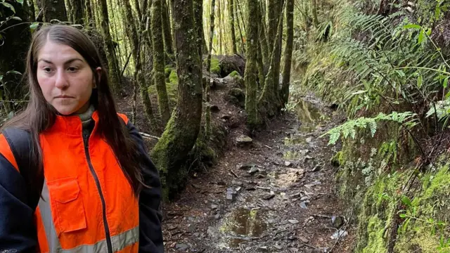 Justine, de pelo largo castaño oscuro, con una chaqueta naranja de alta visibilidad, está de pie en el bosque.