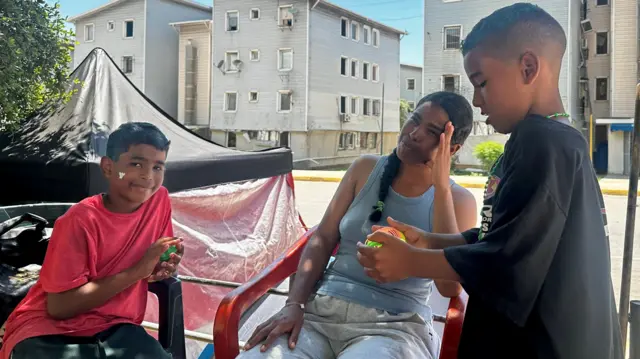 Jhaymar Cantillos sentada en una silla, junto a dos de sus hijos quienes juegan a su alrededor. 