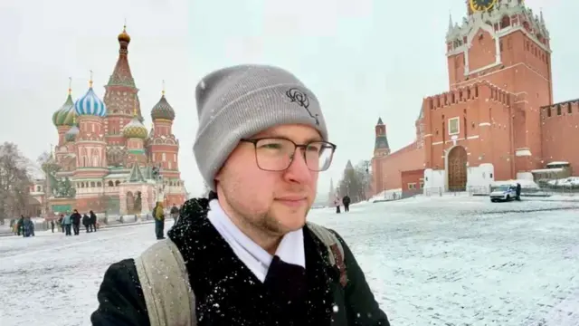 Persona en primer plano que lleva un gorro de lana gris, gafas, un abrigo oscuro y una mochila, con nieve en la ropa y los hombros.
