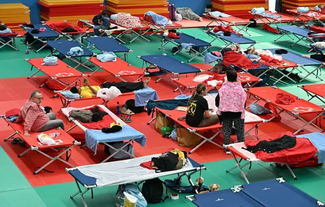 Camillas de campaña de diversos colores puestas en fila en un polideportivo. Hay muchas personas tumbadas o sentadas en sus camillas, viendo el teléfono o conversndo con alguien al lado. 