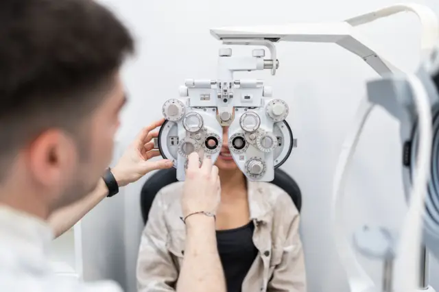 Una mujer está sentada en la silla del oftalmólogo y él le tiene el aparato para probar lentes en frente.