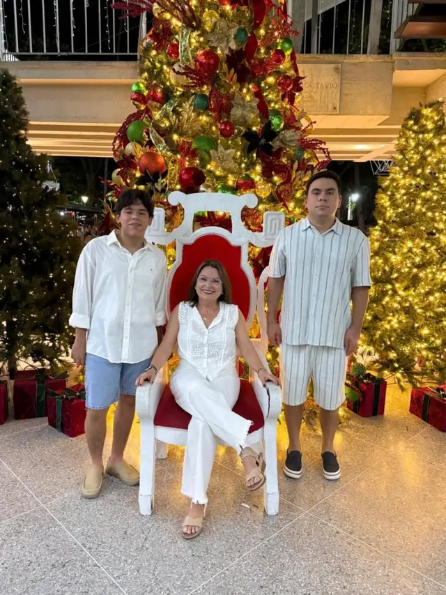 María Lourdes, vestida de blanco y con lentes, está sentada en una silla roja y blanca. A su lado derecho, está parado su hijo Gonzalo, que lleva una camisa blanca y un pantalón corto azul. A su lado izquierdo, está Santiago, que lleva un conjunto blanco con rayas oscuras. Al fondo tienen tres arboles de navidad decorados e iluminados.