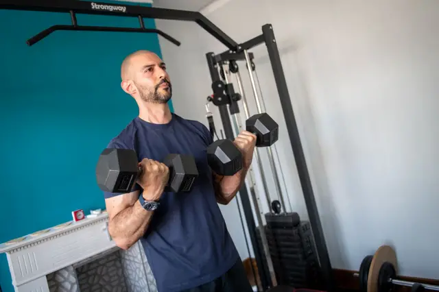 Marios con una camiseta azul oscuro y pantalones cortos haciendo flexiones de bíceps con dos mancuernas.