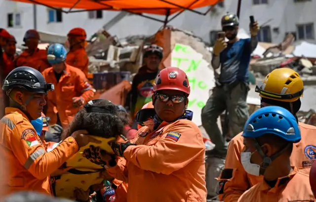 Varios miembros de Protección Civil, vestidos con uniformes anaranjados y cascos. Dos de ellos transportan a una persona en una camilla en el lugar de un edificio derrumbado.