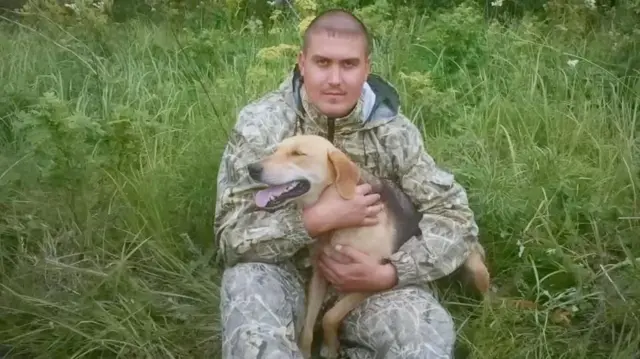 Andrey Spivak, sentado entre hierba alta, sostiene a un perro de color beige y negro. Viste un conjunto de exterior con estampado de camuflaje, lleva el pelo rapado al ras y mira directamente a la cámara.