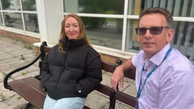 Lowri y el Dr. Healy están sentados en un banco. El Dr. Healy lleva una camisa morada con un cordón, tiene el pelo corto y oscuro y usa gafas. Lowri lleva una chaqueta acolchada negra y jeans.