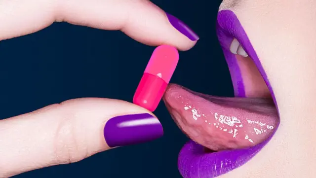 Boca de mujer y dedos de mujer con labios y uñas pintados de púrpura, a punto de tomarse una píldora rosa.