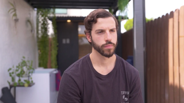 Simon está sentado en su jardín trasero, mirando hacia un lado de la cámara. Tiene barba oscura y cabello corto, ondulado y oscuro. Lleva una camiseta gris oscuro. 
