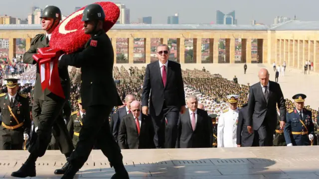 El presidente Recep Tayyip Erdogan sube los escalones del Mausoleo de Atatürk en Ankara durante una ceremonia del Día de la Victoria el 30 de agosto de 2016, flanqueado por funcionarios del gobierno mientras soldados portan una corona de flores con la forma de la bandera turca.