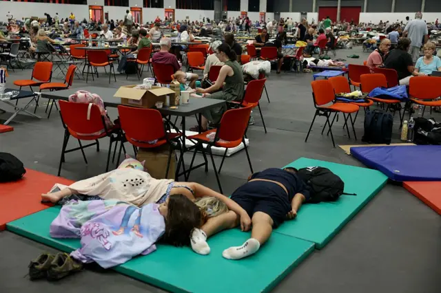 En primer plano, tres personas que parece de la misma familia están echadas sobre colchonetas en el suelo, sin zapatos, tapados con sábanas. Detrás de ellos se ven muchas mesas y sillas donde hay colgadas o puestas mochilas, cajas, enseres varios. Hay infinidad de mesa sy sillas y mucha gente sentada en ellas. 