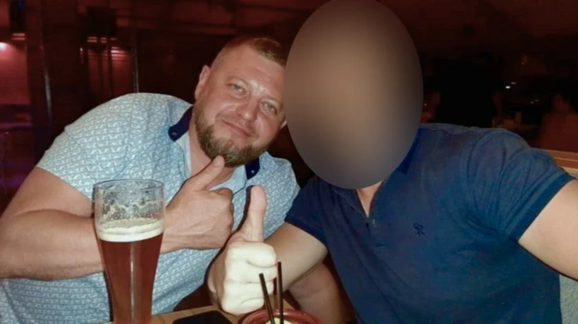 Dos hombres, con vasos grandes frente a ellos, hacen el gesto de pulgar hacia arriba a la cámara. El de la izquierda, Ruslan Yeriomichev, lleva barba recortada y el pelo muy corto, y viste una camiseta clara con estampado. El rostro del hombre de la derecha ha sido difuminado.