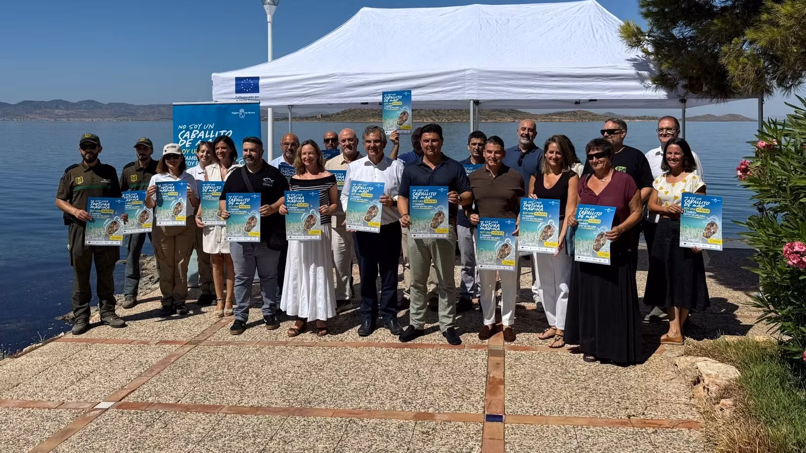 Autoridades y asistentes sostienen carteles de la campaña de sensibilización sobre la nacra durante el acto celebrado en La Manga del Mar Menor.