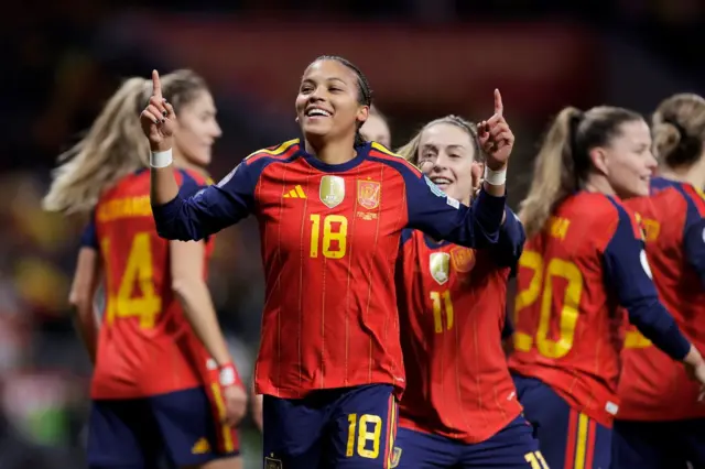 Vicky López, vestida con el uniforme de la selección española, sonríe mientras sus dedos índices apuntan hacia el cielo. En el fondo se ven otras futbolistas españolas