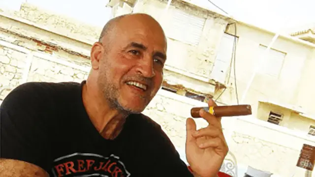 Daniel Siad, un hombre con la cabeza rapada y barba incipiente, que viste una camiseta negra, está sentado al aire libre en lo que parece ser una calle soleada, sonriendo con un gran puro en la mano.