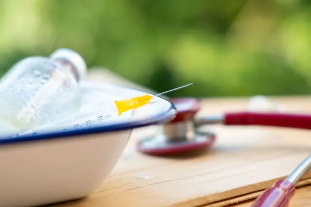 Un plato con una jeringa y un vial en una mesa, y un estetoscopio detrás fuera de foco. 