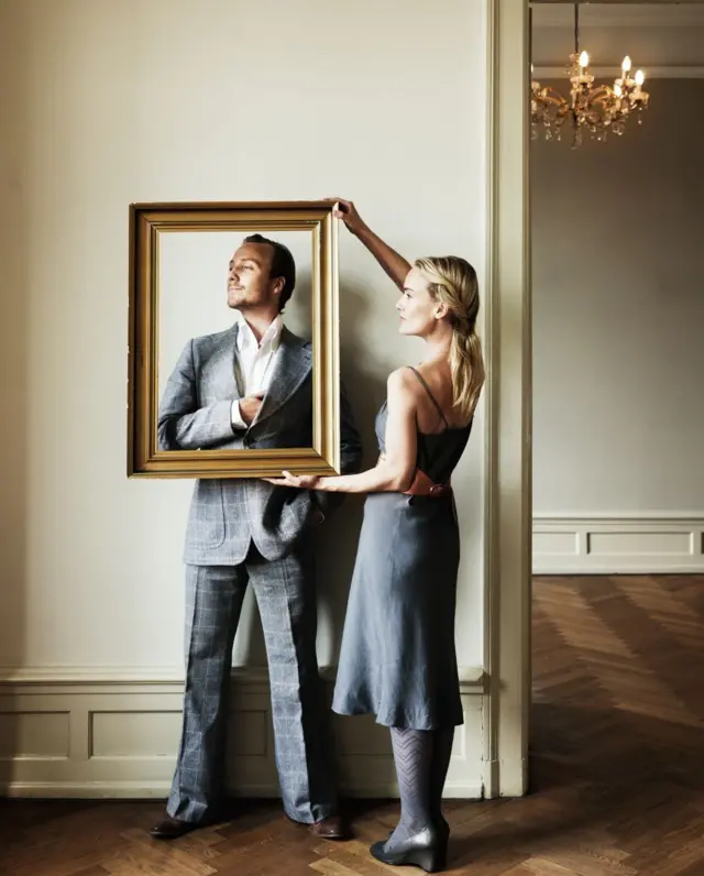 Mujer vestida de gris le pone un marco dorado a un hombre que está recostado a una pared, posando, en un apartamento elegante.