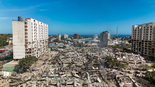 Dos edificios, gravemente afectados, están de pie. En el centro, se ven edificaciones totalmente colapsadas. Grupos de personas caminan en el área. El cielo azul está totalmente despejado.